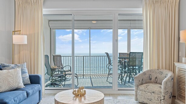 A living room with a blue sofa, a round wooden coffee table, and a patterned armchair. A glass door leads to a balcony with a view of the ocean.