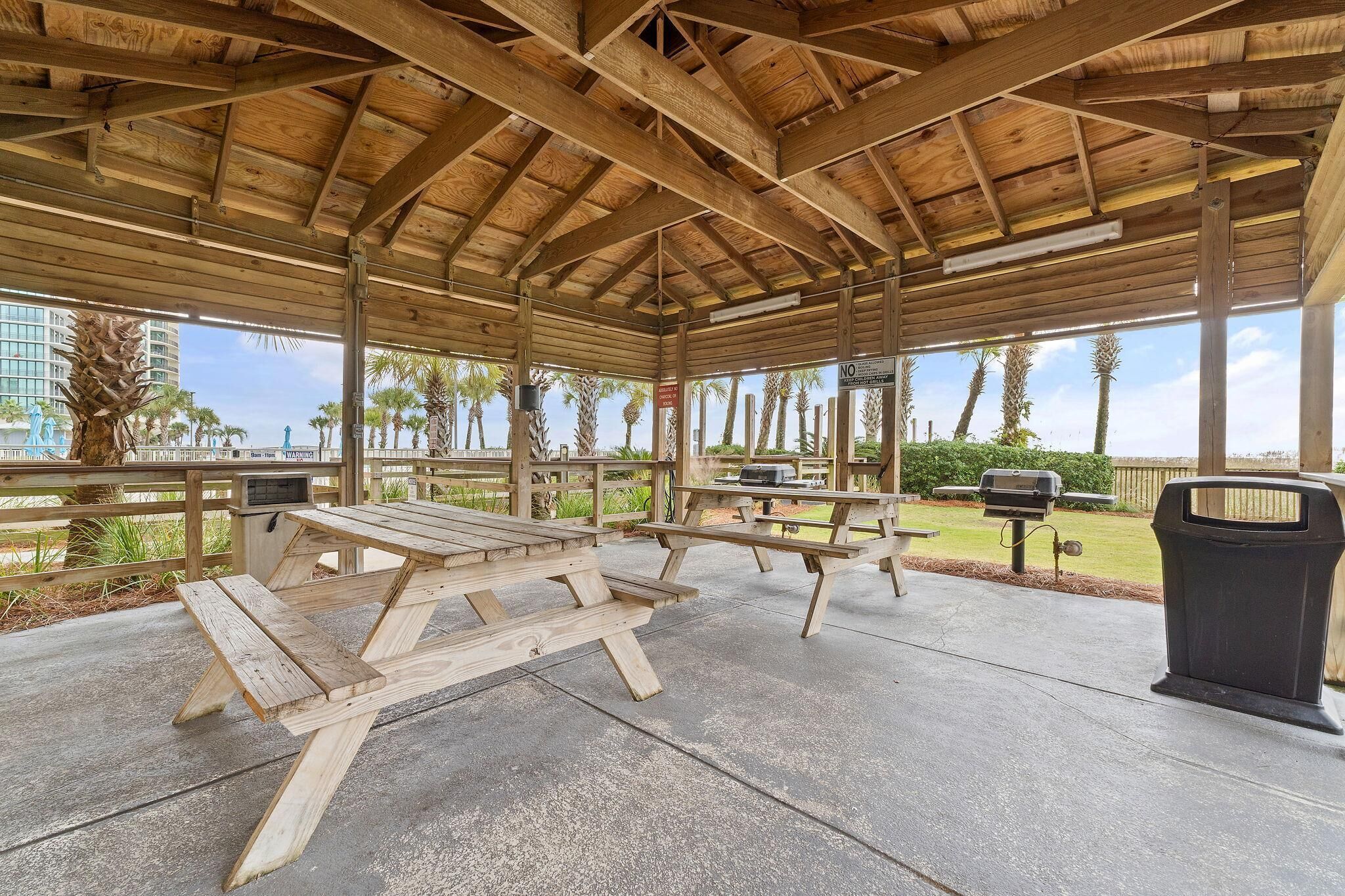 A covered picnic area with wooden benches and picnic tables, a grill, and a trash can.