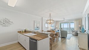 A spacious kitchen with a granite countertop, white cabinets, and a dishwasher. There is a dining area with a table and chairs, and a view of the ocean through large windows.