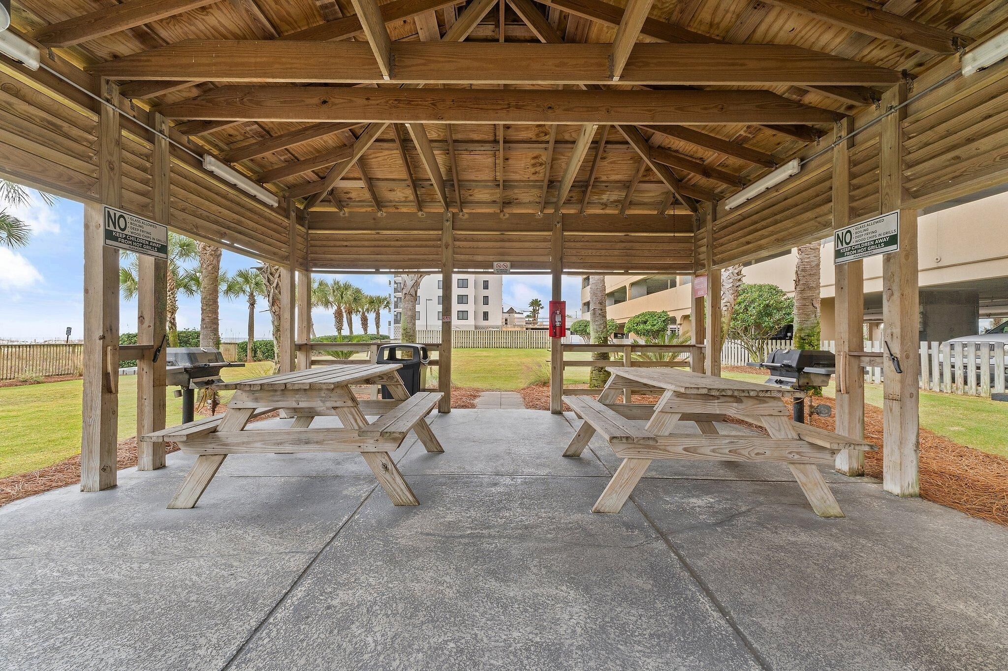 A covered picnic area with wooden benches and picnic tables, a grill, and a view of a building in the background.