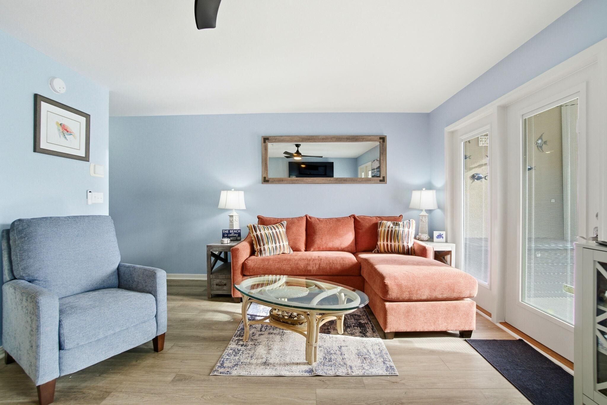 A living room with a blue armchair, a peach-colored sofa, a glass coffee table, and a mirror on the wall.