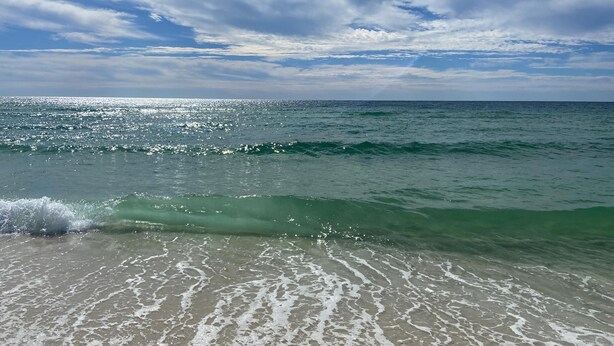 A beach with clear water and waves, under a partly cloudy sky.