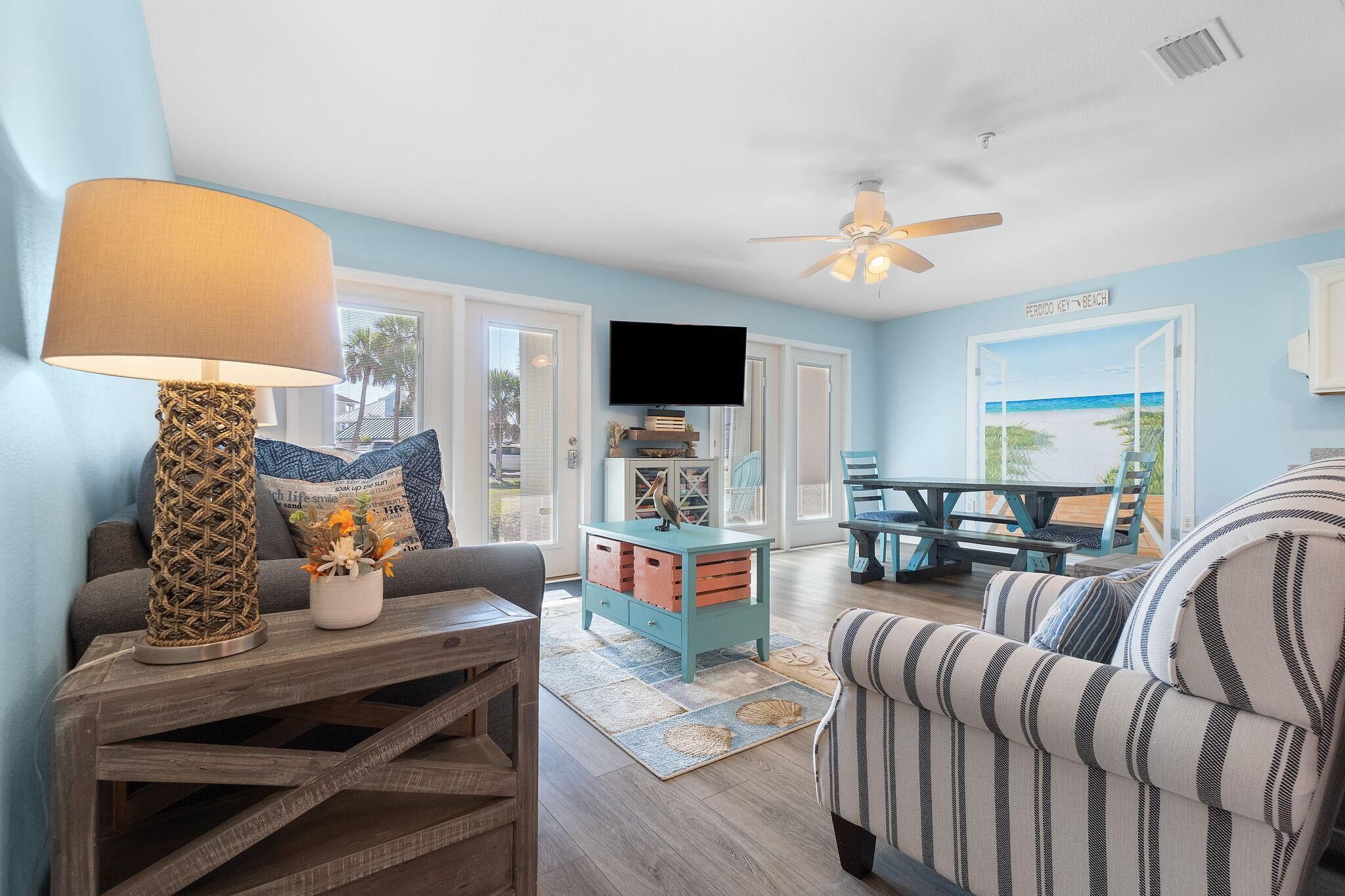 A living room with a TV, a wooden coffee table, a striped armchair, and a view of the beach through an open door.