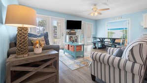 A living room with a TV, a wooden coffee table, a striped armchair, and a view of the beach through an open door.