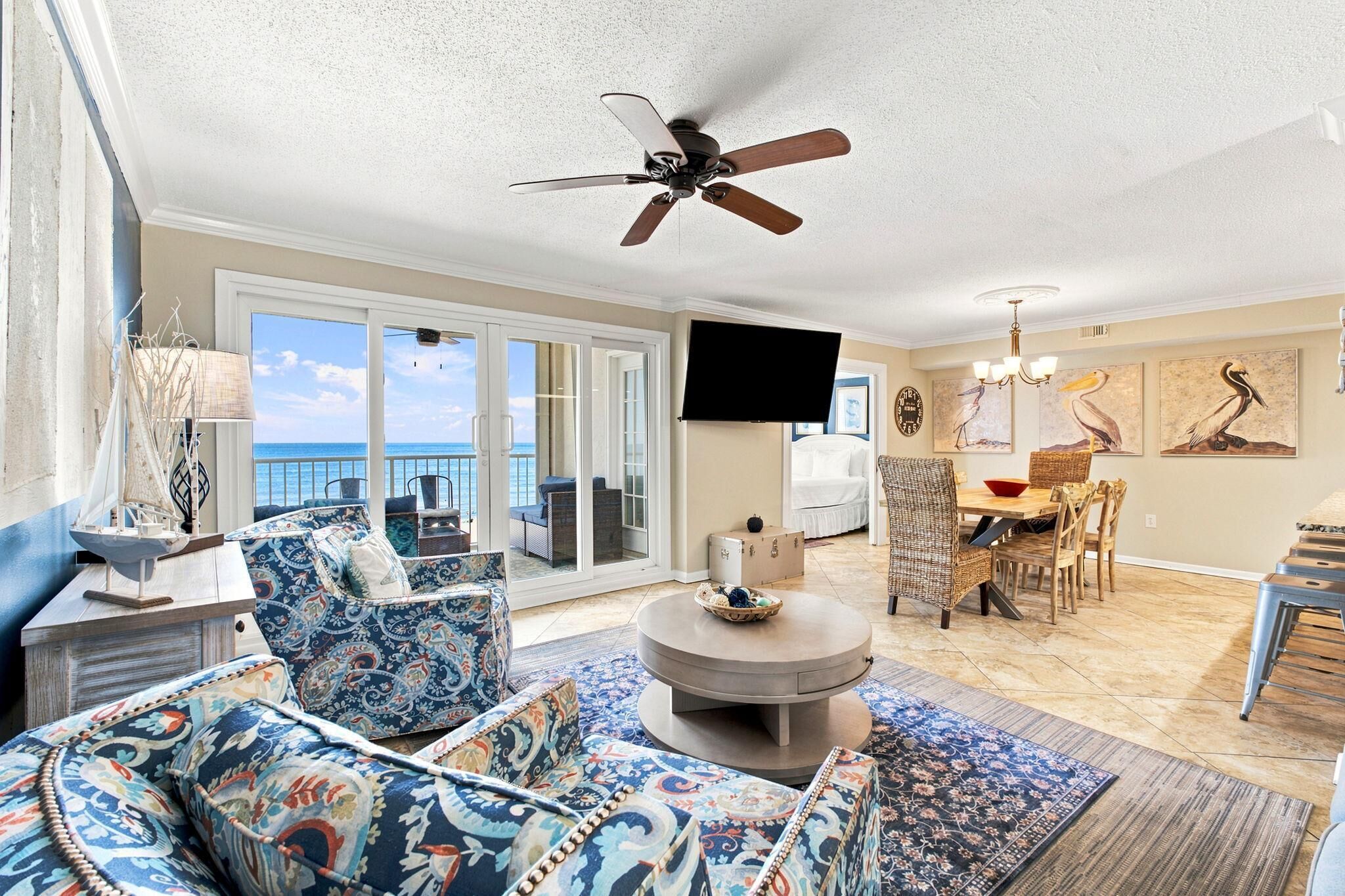 A spacious living area with a ceiling fan, a dining table with chairs, and a view of the ocean through large sliding doors.