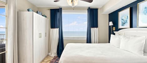 Habitación de hotel con cama, ventilador de techo, ventana con cortinas y vista al mar.