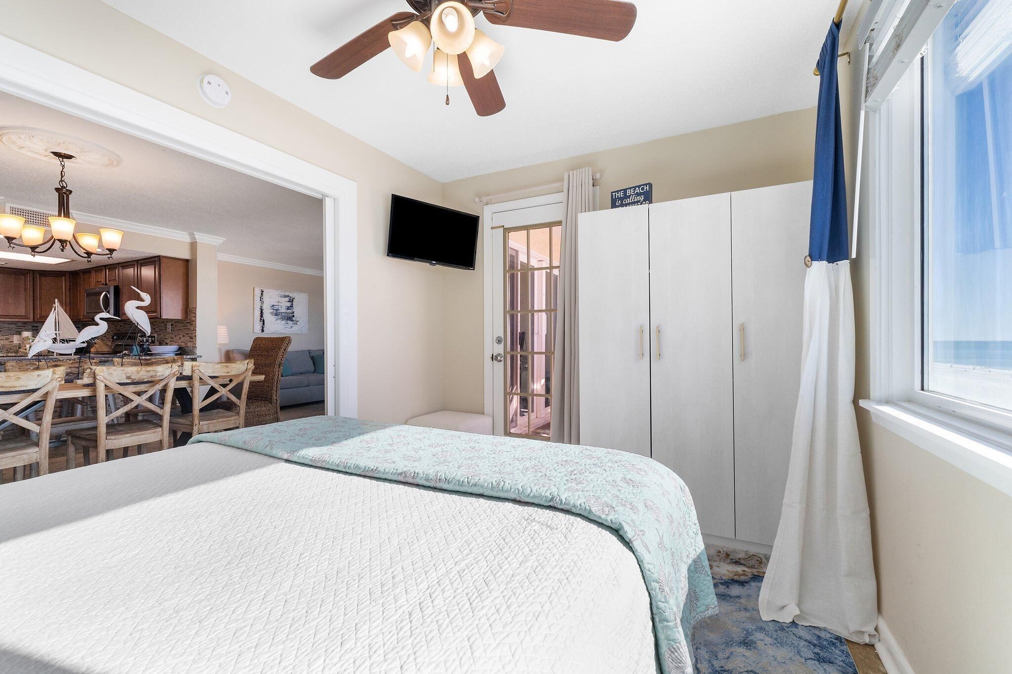 A bedroom with a bed, a ceiling fan, a TV, a desk with chairs, and a view of the beach.