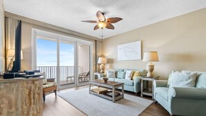A spacious living room with a large window, a ceiling fan, a sofa, a coffee table, and a side table with lamps. - Windemere 602 (Pensacola)