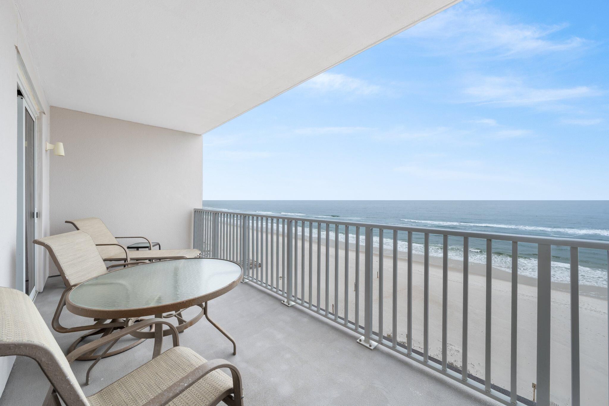 A balcony with a glass table and chairs, overlooking a beach with a railing.