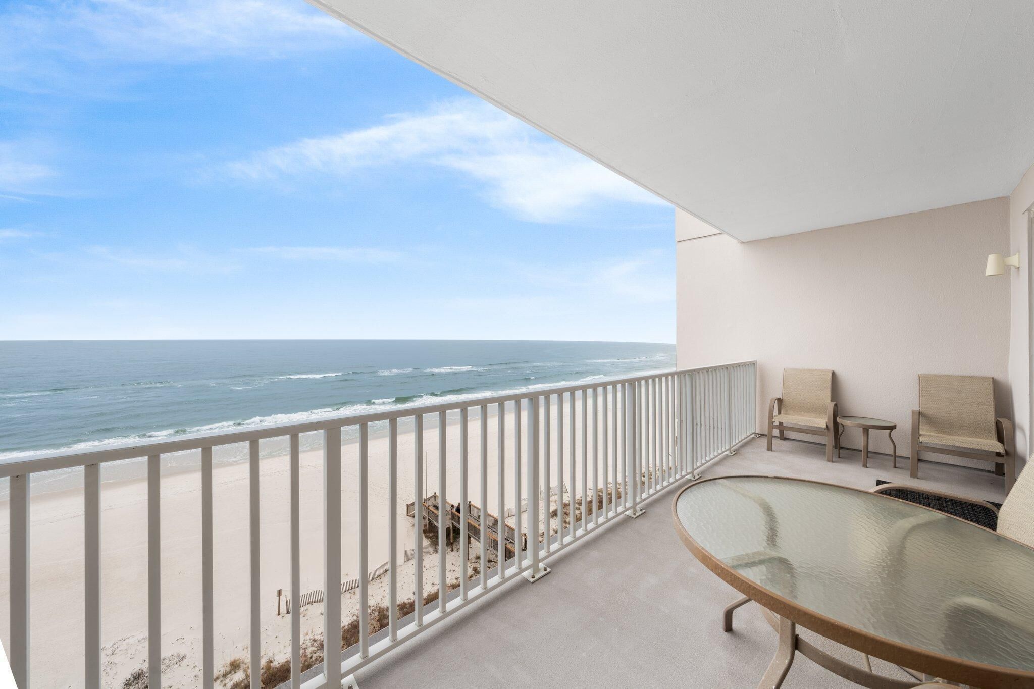 Een balkon met een glazen tafel en twee stoelen, met uitzicht op een strand met een balustrade.