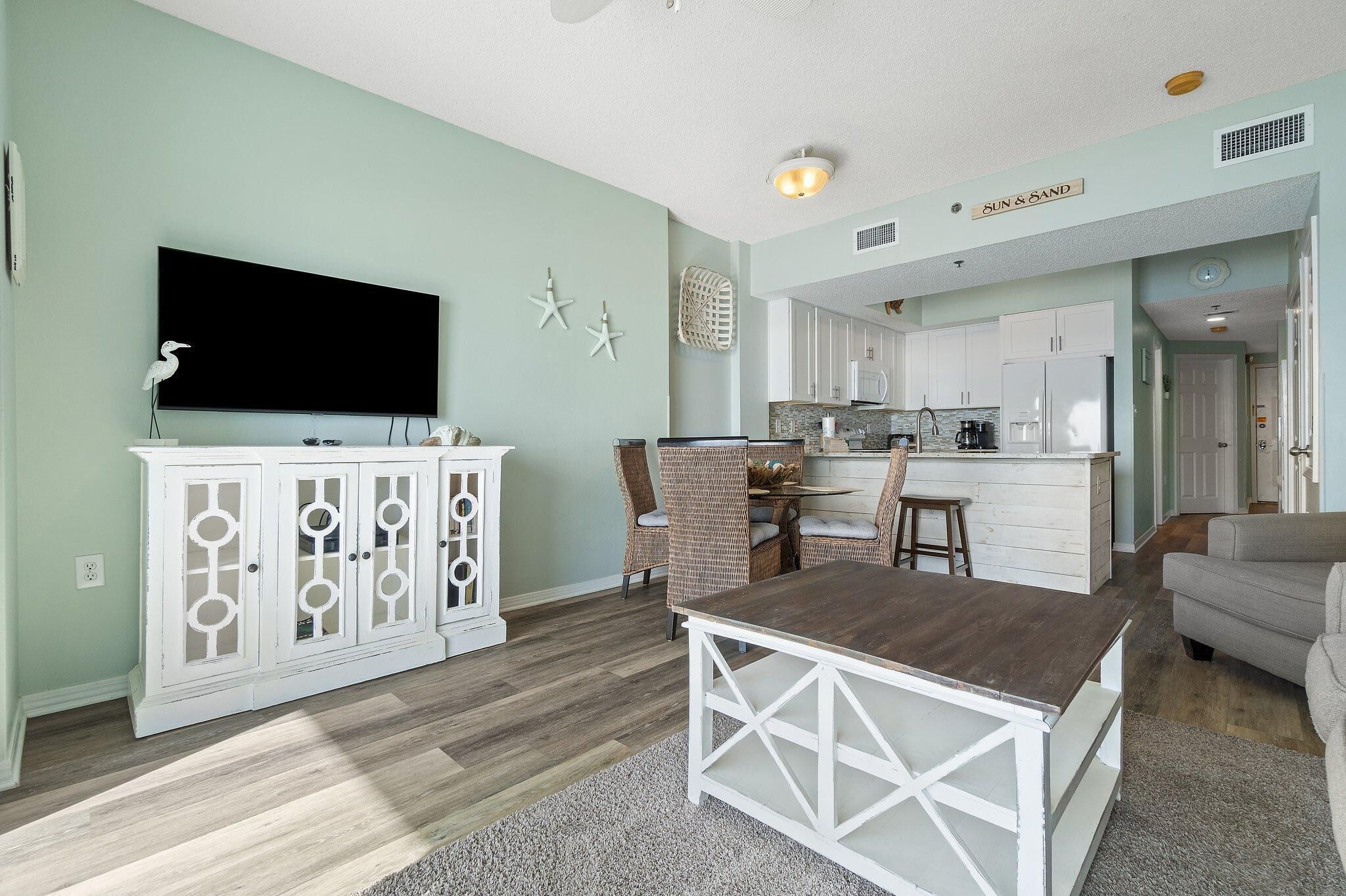 A living room with a TV, a dining table, and a kitchen in the background.