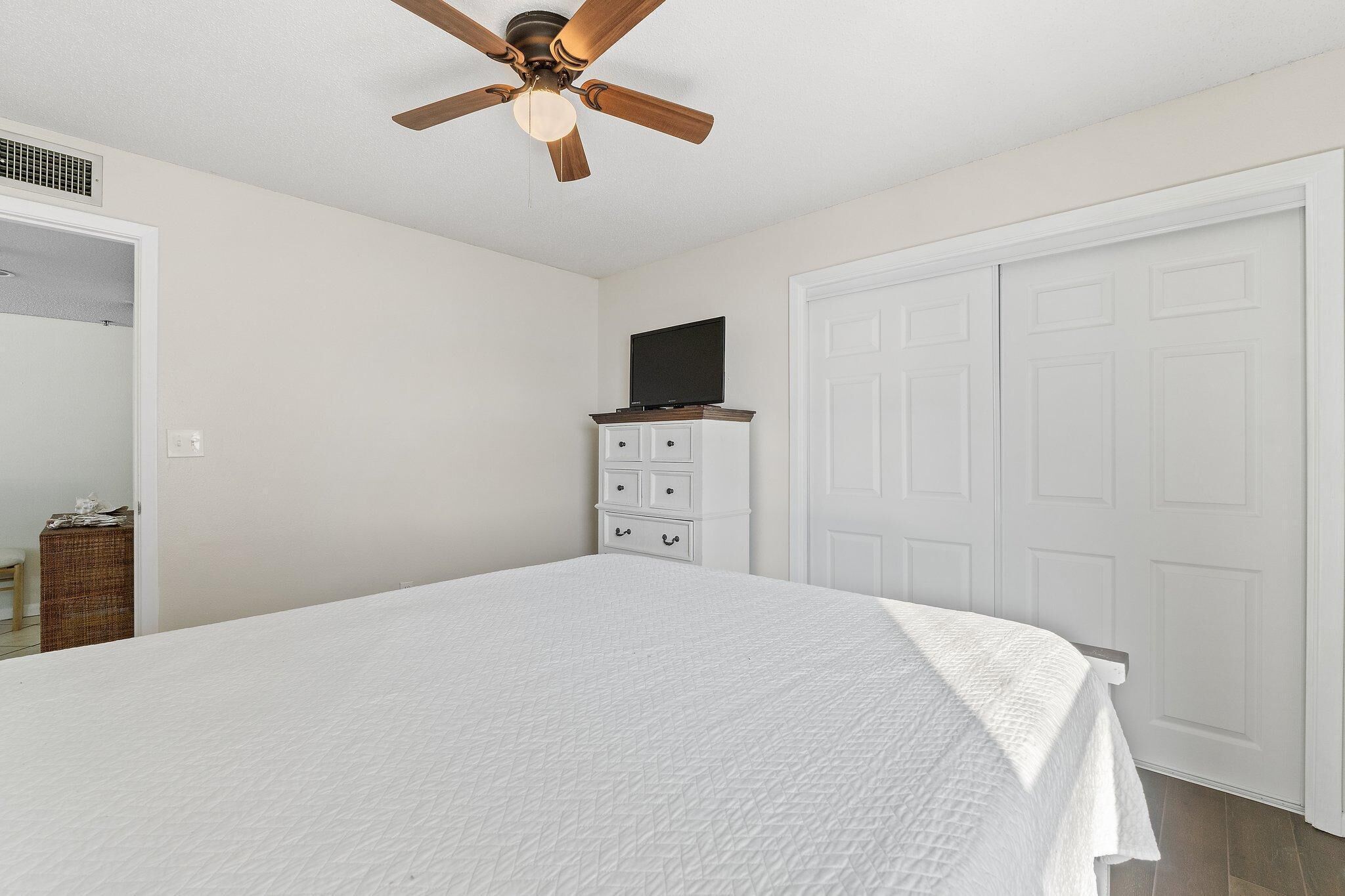 A bedroom with a bed, a ceiling fan, a dresser, a television, and a white door.