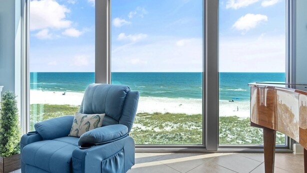 Habitació amb una finestra gran que dona a una platja, un butac blau i un piano.
