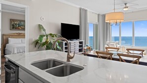 A modern hotel room with a large island sink, a flat-screen TV, a dining area with wooden chairs, and a view of the ocean. - Regency Isle Penthouse 1106 (Orange Beach)
