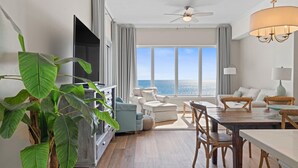 A modern living room with a large window, a ceiling fan, and a dining area with wooden furniture. - Regency Isle Penthouse 1106 (Orange Beach)