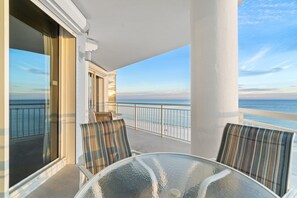Un balcon avec une table en verre et des chaises à rayures, donnant sur une plage protégée par une rambarde.