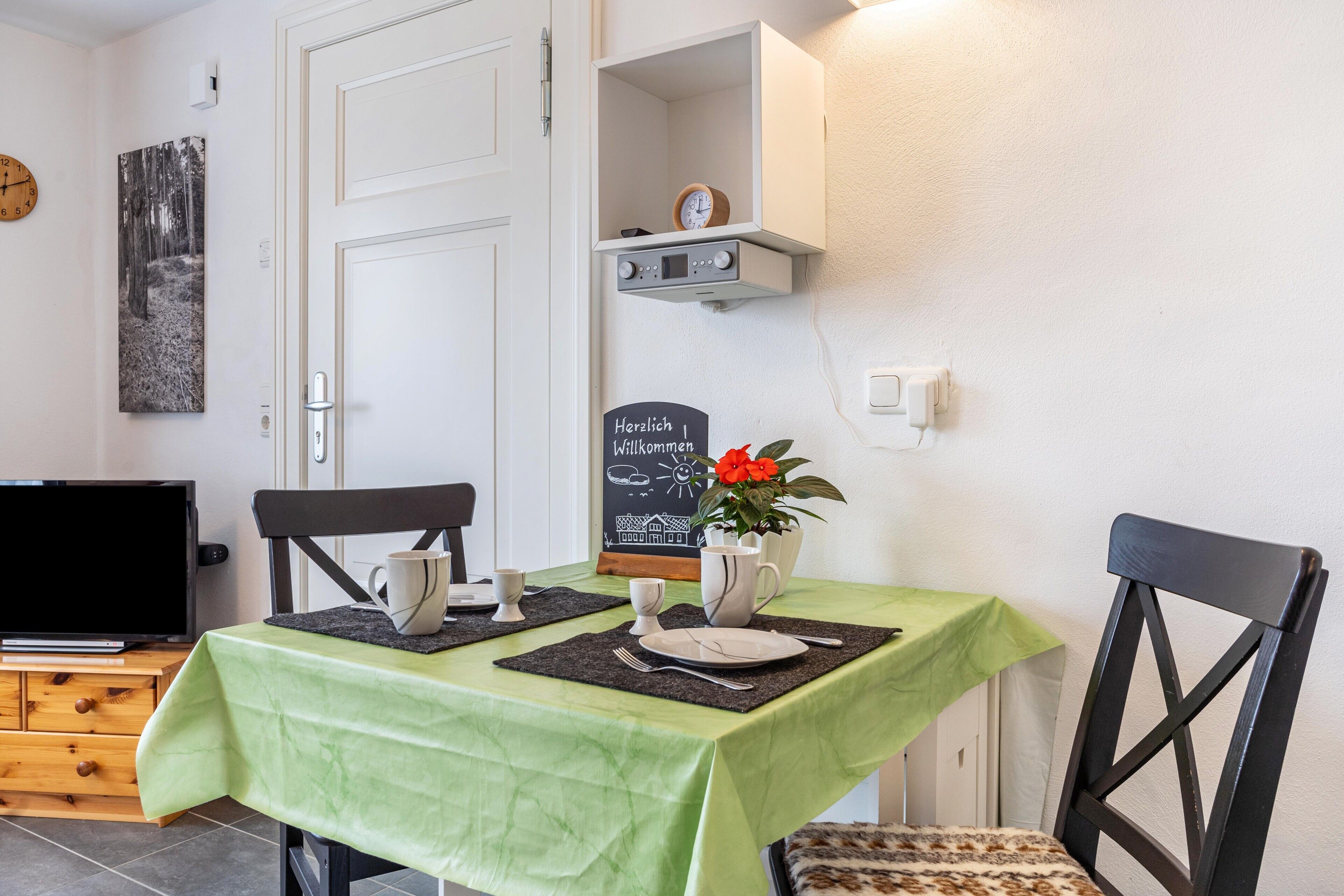 Un comedor con mantel verde, sillas, televisor, reloj y letrero de bienvenida.