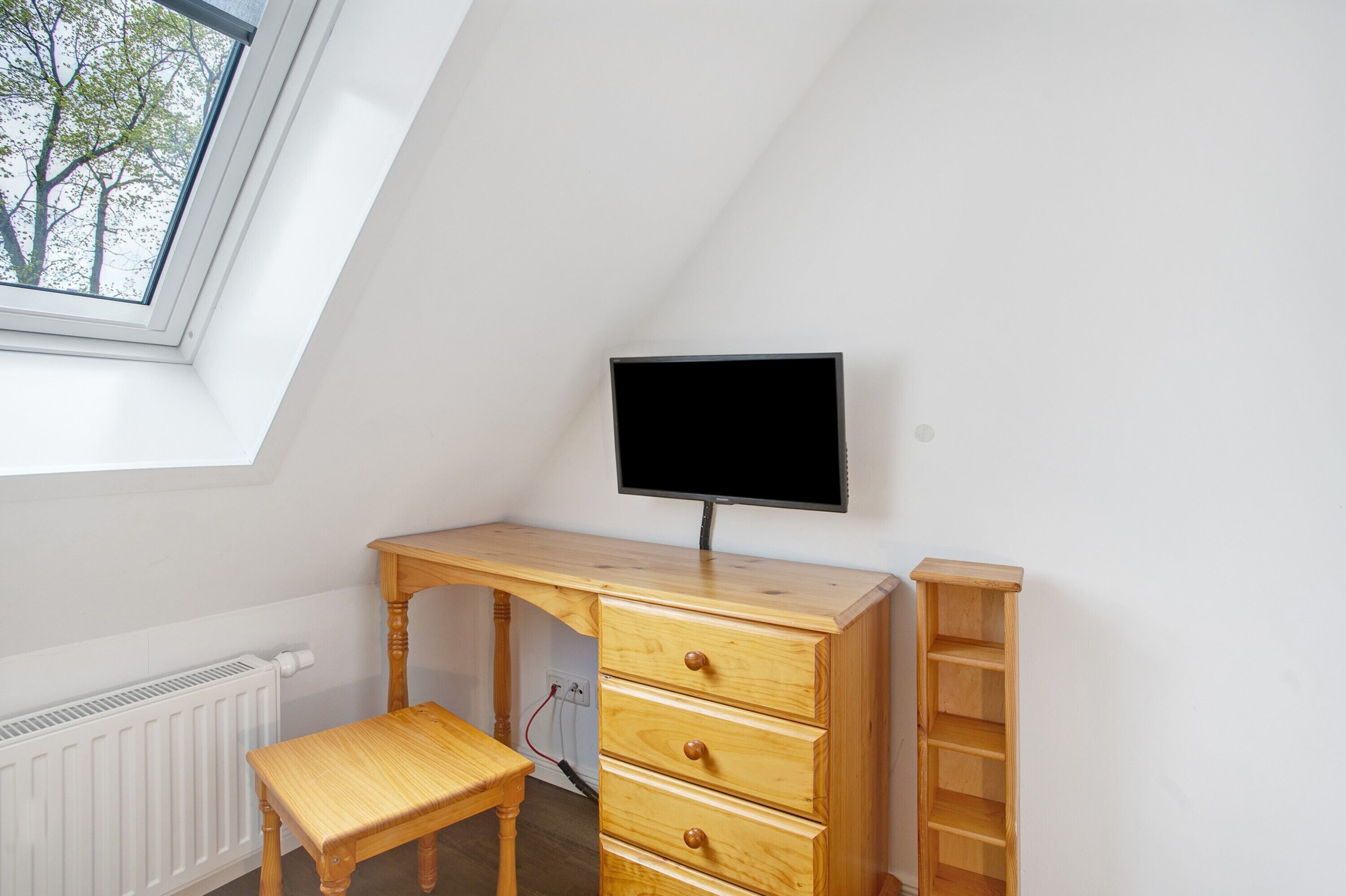 Una stanza con soffitto a falde, una scrivania in legno con cassetti, un televisore, un termosifone e una finestra vista alberi.