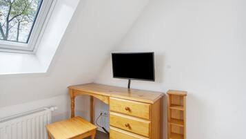 Una stanza con soffitto a falde, una scrivania in legno con cassetti, un televisore, un termosifone e una finestra vista alberi.