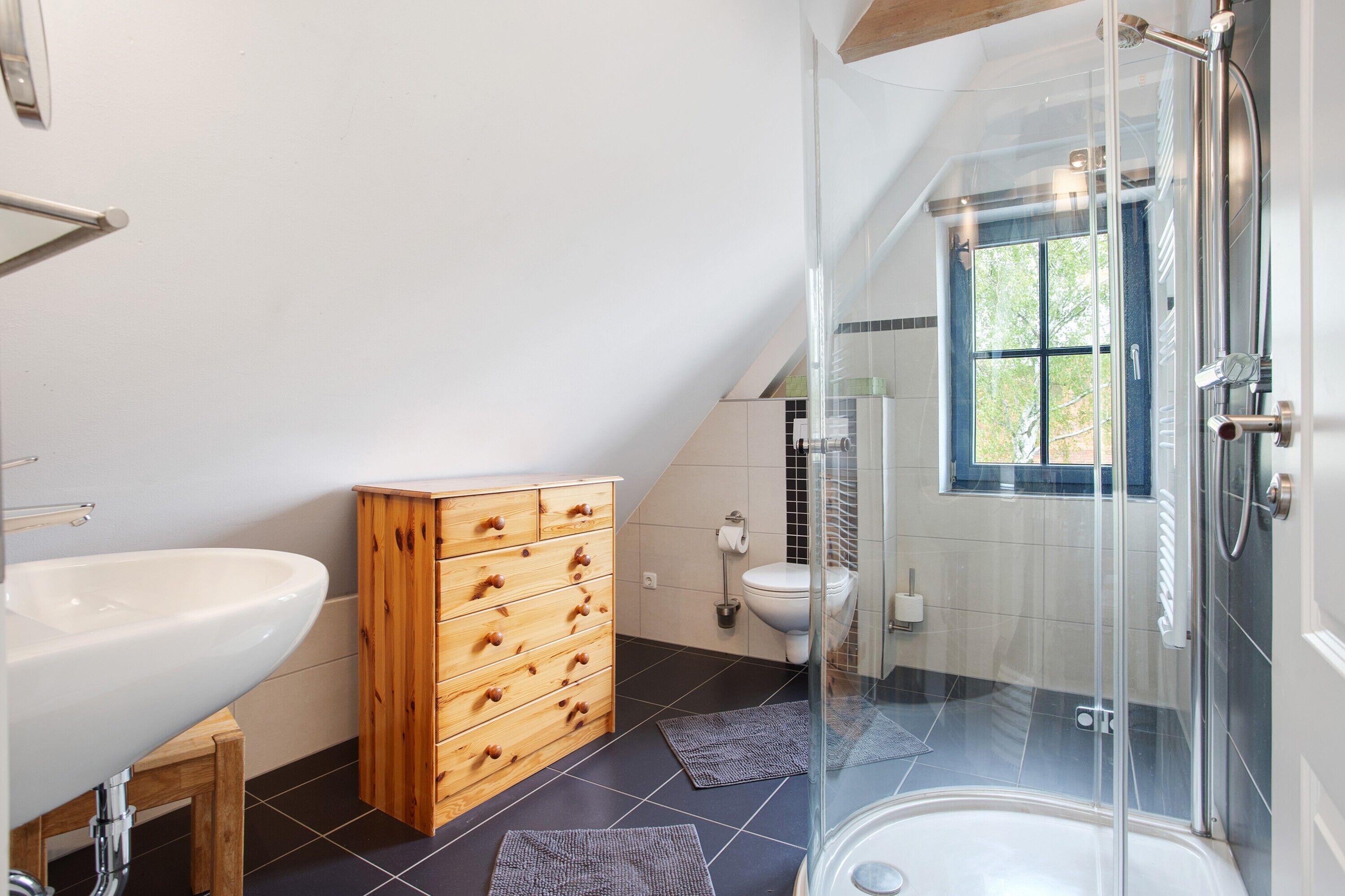 Et moderne badeværelse med en glasvæg omkring brusebad, et træskab, en hvid vask og et vindue med udsigt.