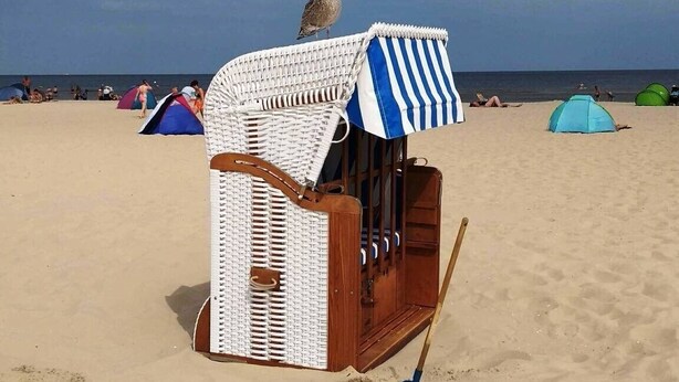 Een zandstrand met een wit- en blauw gestreepte strandparasol, een rieten strandstoel en een blauwe schep in het zand.
