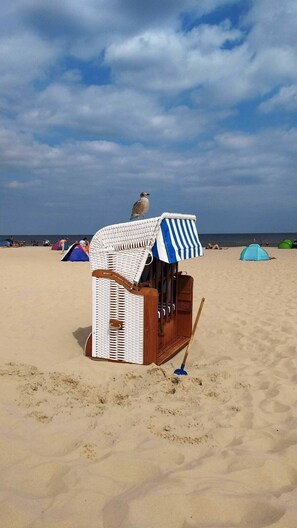 A beach scene with a white and blue striped beach umbrella, a wicker beach chair, and a blue shovel on the sand. - Apartment 'Kornkammer' with Lake View, Private Garden and Wi-Fi (Usedom)