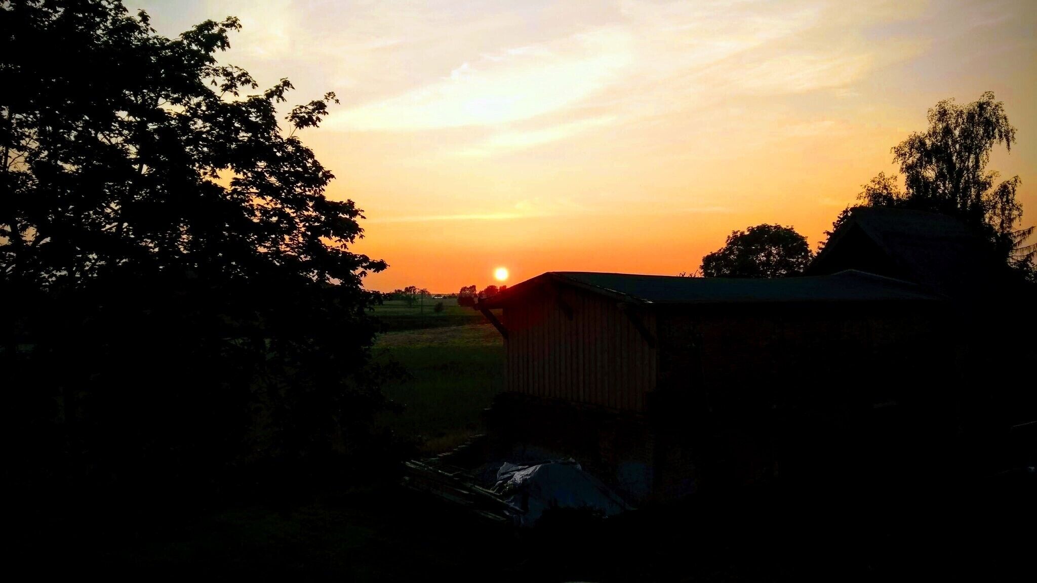 A sunset over a rural landscape with a barn and trees.
