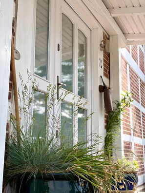 Eine weiße Tür mit Fenster, ein Topf mit hohem Gras und eine Ziegelwand.