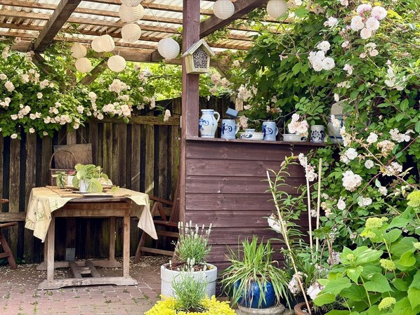 Eine rustikale Außenanlage mit einem Holztisch unter einer Pergola, umgeben von üppigem Grün und blühenden Pflanzen.