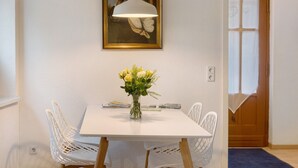 Uma área de refeições com uma mesa branca, cadeiras, um vaso com flores, uma luminária pendente e um quadro na parede.