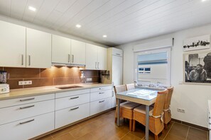 Une cuisine moderne avec des meubles blancs, une table à manger entourée de chaises en osier et une fenêtre avec des stores.