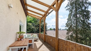 Een houten terras met een tafel en stoelen, een overdekt gedeelte en uitzicht op bomen en gebouwen.