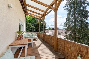 Una terraza de madera con una mesa y sillas, un espacio cubierto y vistas a árboles y edificios.