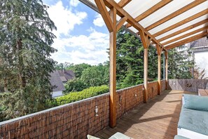 Une terrasse en bois agrémentée d’une pergola, d’un espace salon extérieur et offrant une vue sur les arbres et les maisons.