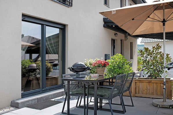 Une terrasse extérieure avec une table et des chaises, un barbecue et un grand parasol.