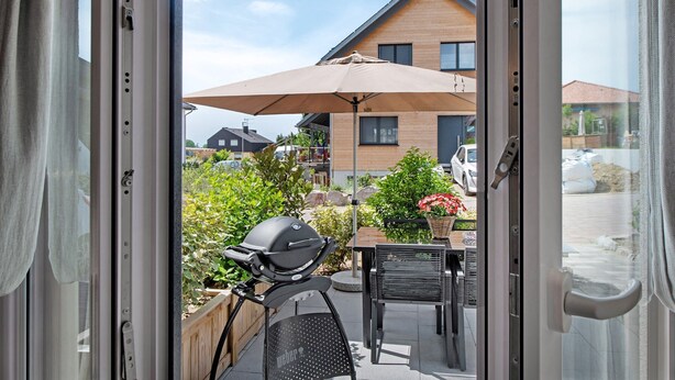 Vista a través de una puerta corrediza de vidrio hacia un patio al aire libre con una parrilla, una mesa y una sombrilla.