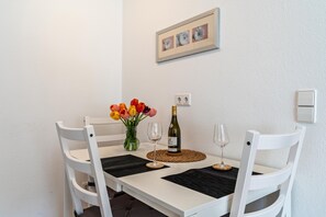 Une table à manger blanche avec des sets de table noirs, deux chaises blanches, une bouteille de vin, deux verres à vin et un vase de tulipes.