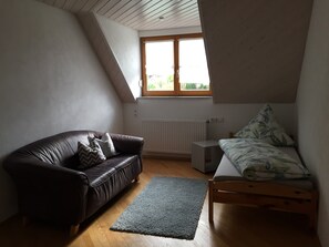 A loft bedroom with a sloped ceiling, a window with a view, a brown leather sofa, a bed with a patterned blanket, and a small bedside table.