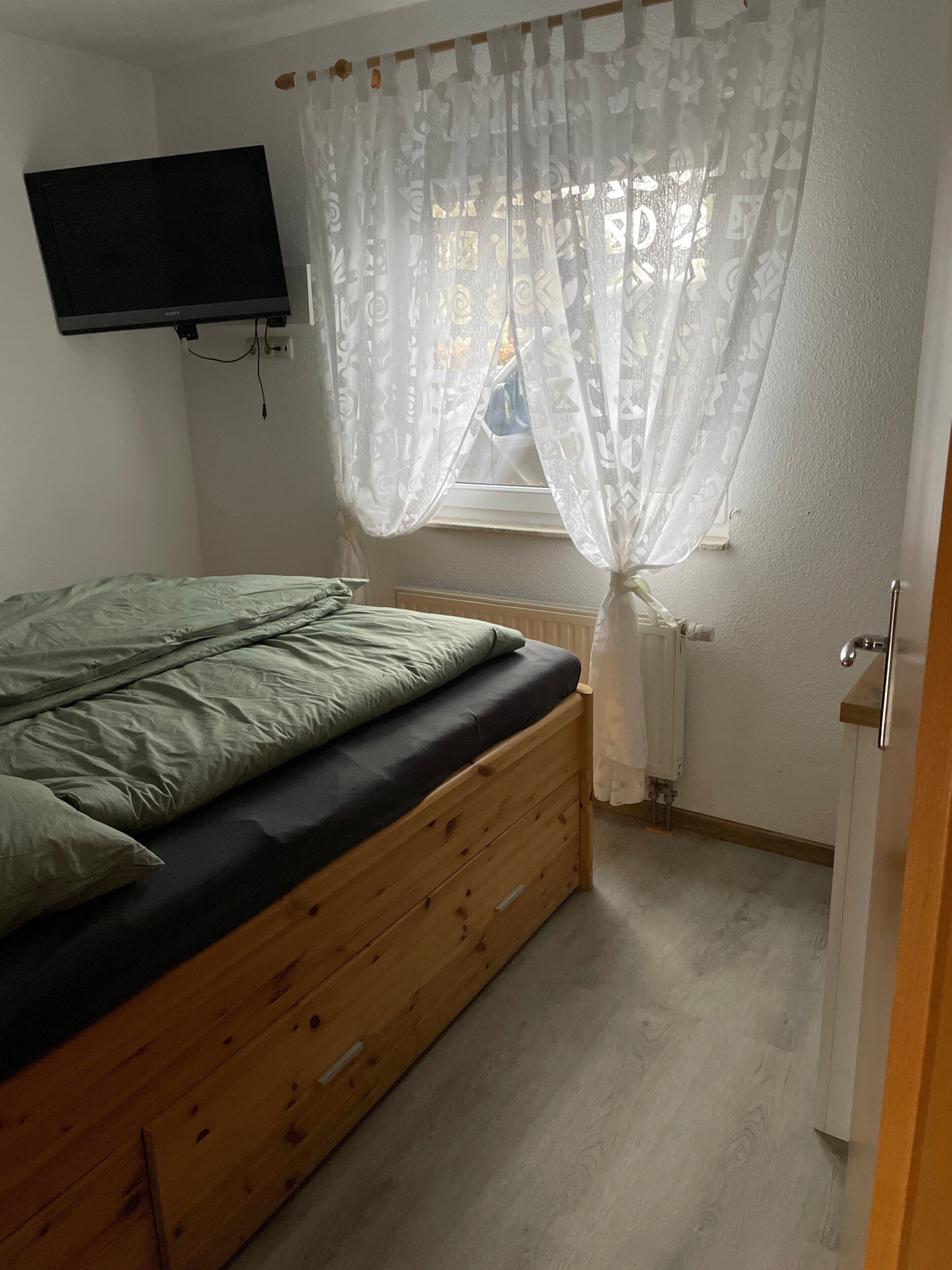 A bedroom with a bed, a television mounted on the wall, a window with sheer curtains, and a radiator.