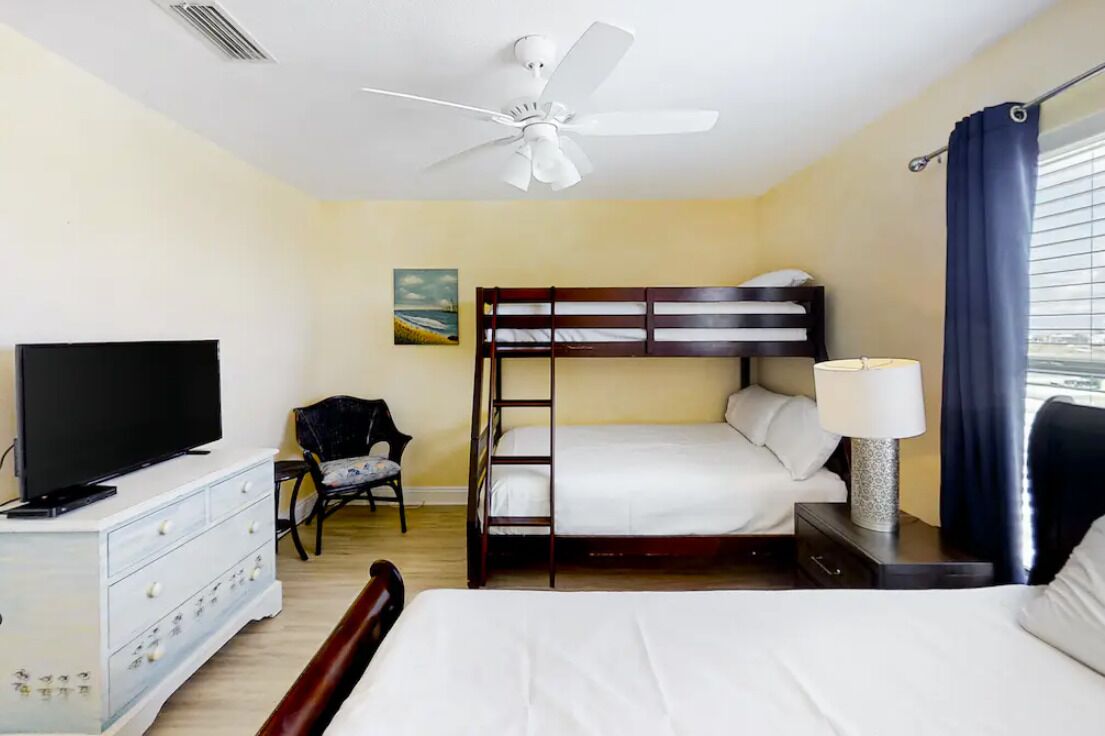 Une chambre d’hôtel avec un lit superposé, un téléviseur posé sur une commode, un ventilateur de plafond et une fenêtre avec des rideaux.