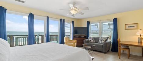 A hotel room with a large bed, a ceiling fan, a TV, a sofa, a dining table, and a view of the ocean.
