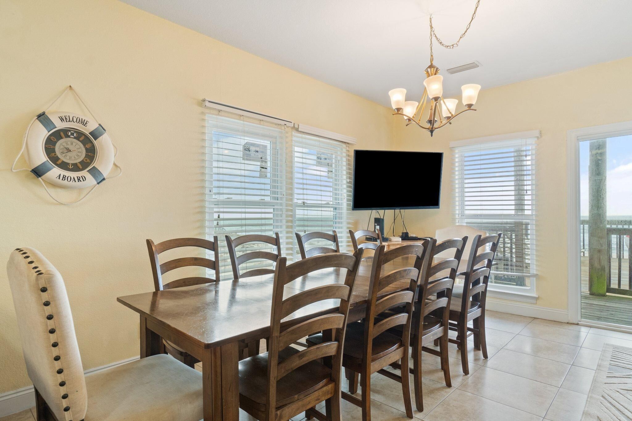 A dining area with a wooden table and chairs, a life preserver wall decoration, a flat-screen TV, and a view of the outdoors through large windows.