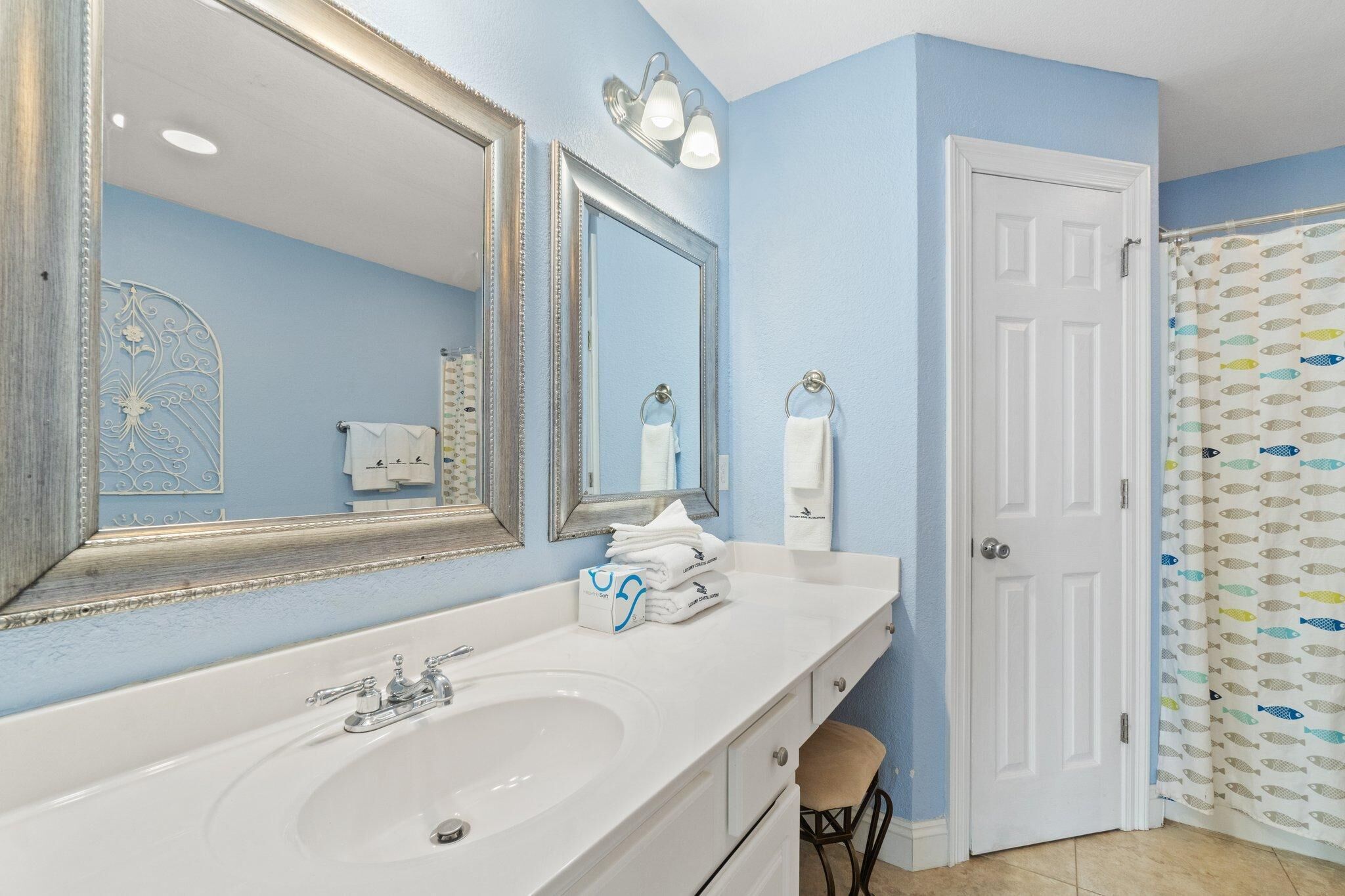 Une salle de bain avec un lavabo blanc, deux miroirs, un porte-serviettes et un rideau de douche orné de motifs de poissons.