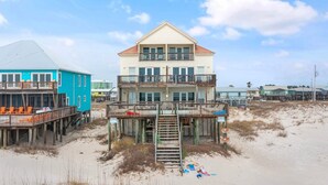 Une propriété située en bord de mer, comprenant une maison à deux étages dotée d’un balcon, d’une terrasse en bois et d’une plage de sable.