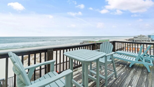 Une terrasse en bois avec des fauteuils Adirondack bleu clair et une petite table, donnant sur une plage sous un ciel dégagé.