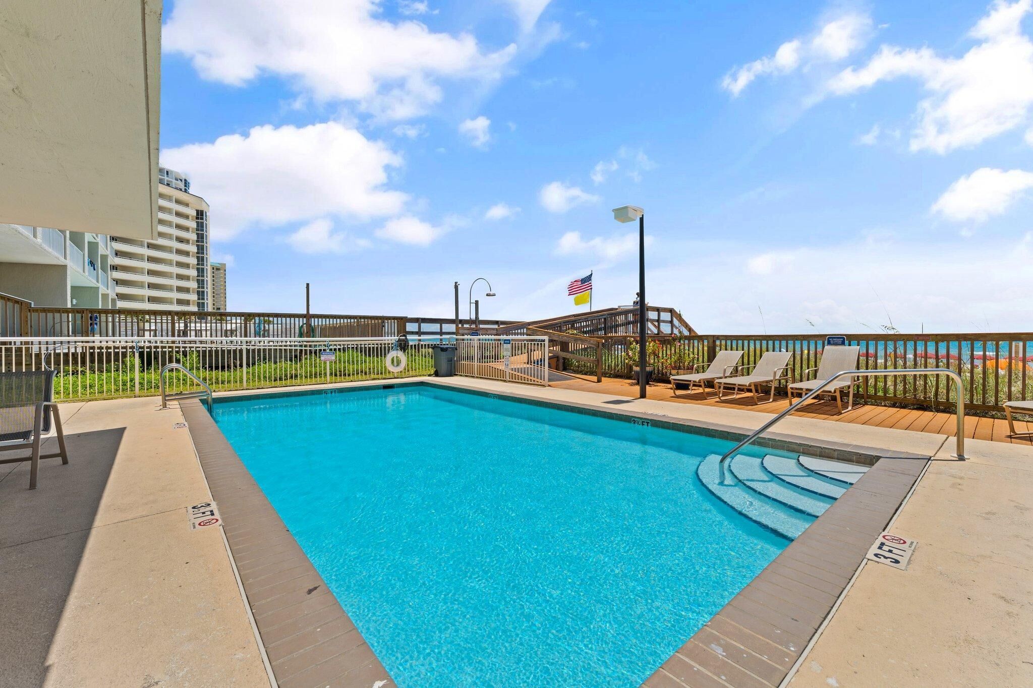 A rectangular swimming pool with a wooden deck, lounge chairs, and a flag.