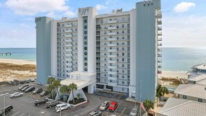 A multi-story hotel with a central atrium, parking lot, and a view of the beach and ocean. - Summerchase 506 (Orange Beach)