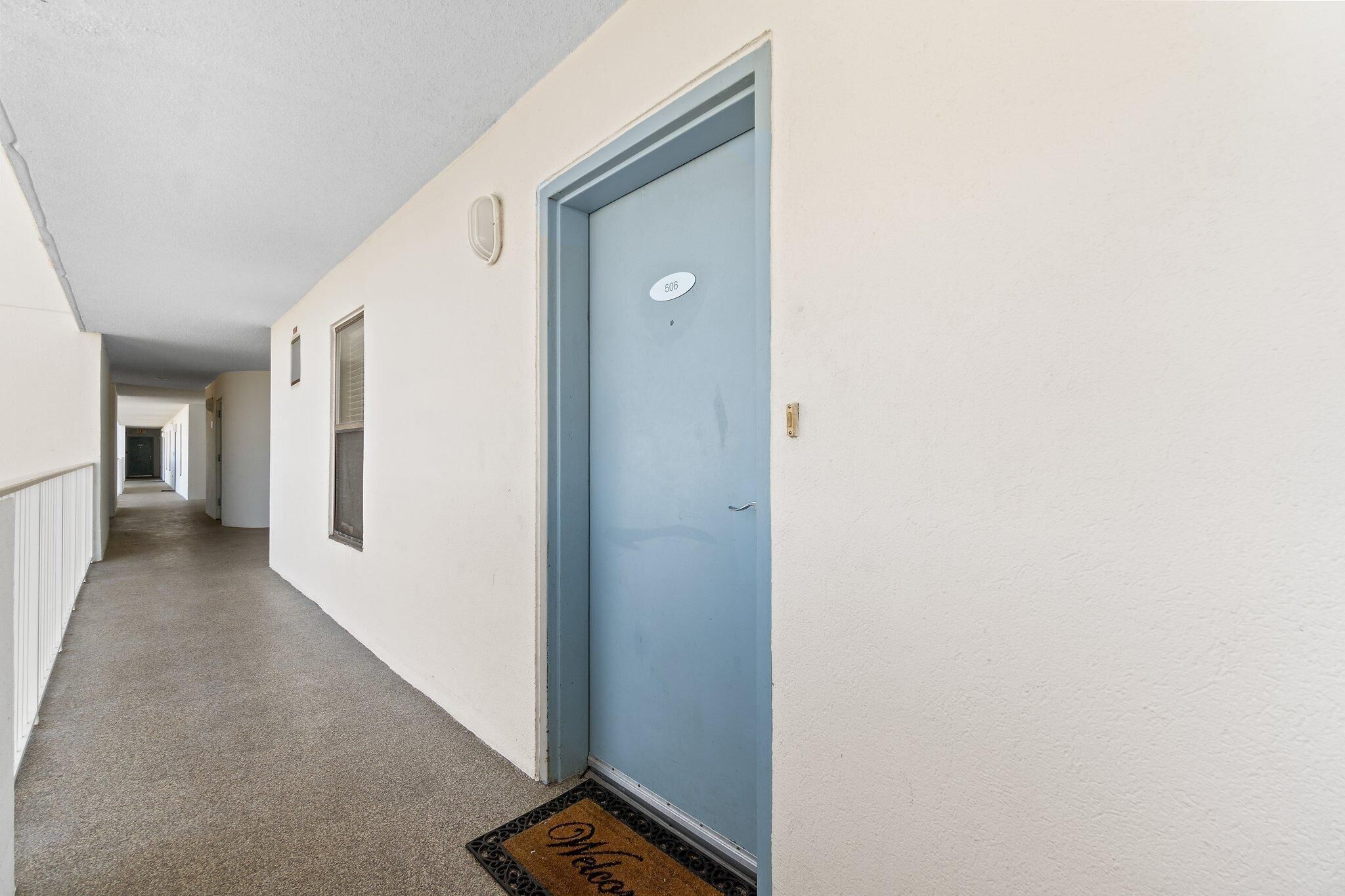 A hallway with a blue door marked with the number 506, a doormat, and a light switch on the wall.