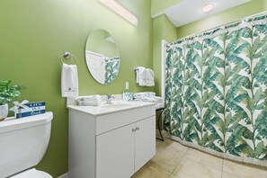 Une salle de bain avec un mur vert, un meuble-lavabo blanc, un miroir, un rideau de douche orné de motifs de feuilles tropicales et une pancarte sur laquelle on peut lire : « LA VIE EST UNE PLAGE ».