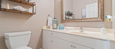 A bathroom with a white toilet, a vanity with a mirror, and a shelf with plants and toiletries.